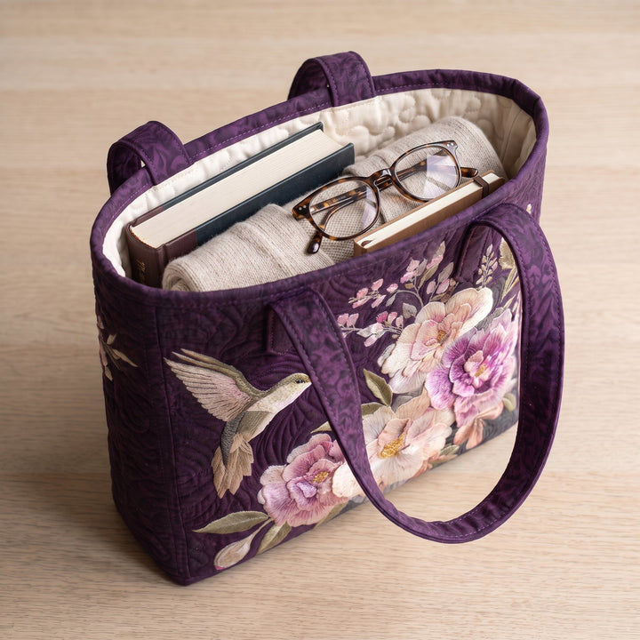 Bolso Artesanal Acolchado — Colibríes y Orquídeas: con un libro de tapa dura, un suéter doblado y lentes de lectura